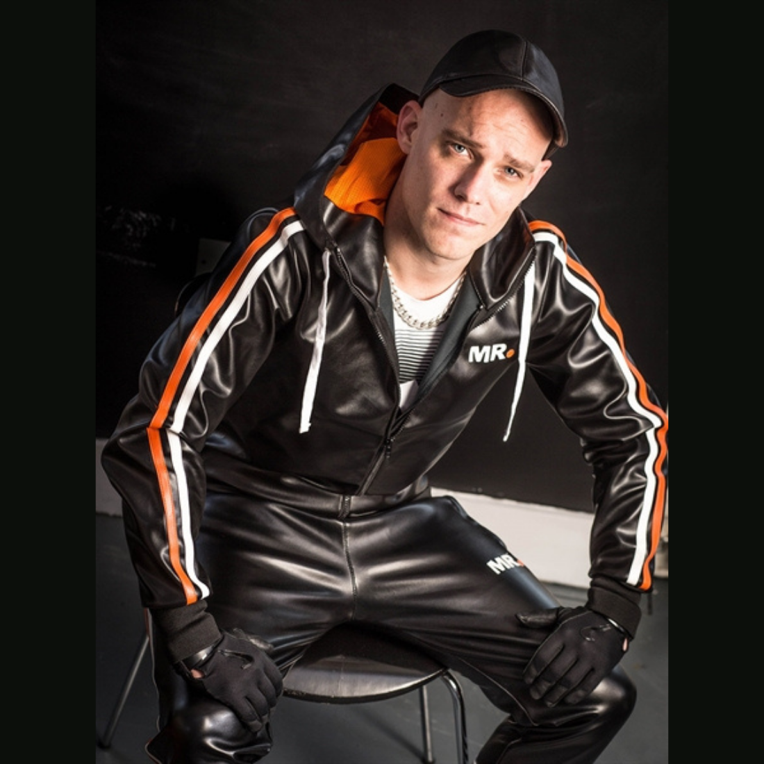 Zwarte kunstleren trainingsjack met oranje en witte strepen, ontworpen door Mr. Riegillio. Perfect voor gay mannen en te koop bij Flavourez.