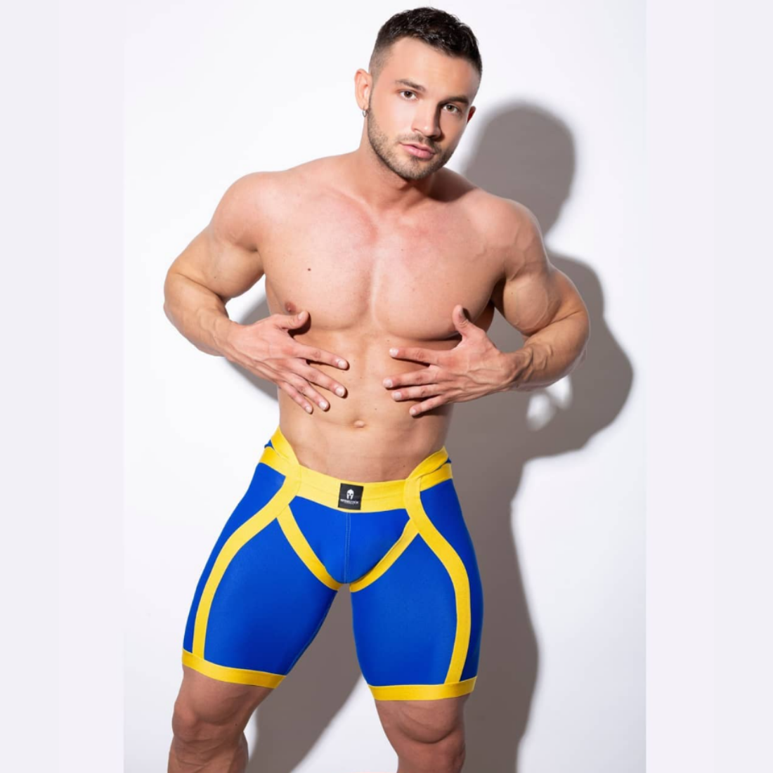 Unieke blauwe short met verlaagde tailleband aan de voorzijde en gele elastische banden, ontworpen door het Italiaanse modehuis Sparta’s Harness en te koop bij Flavourez.